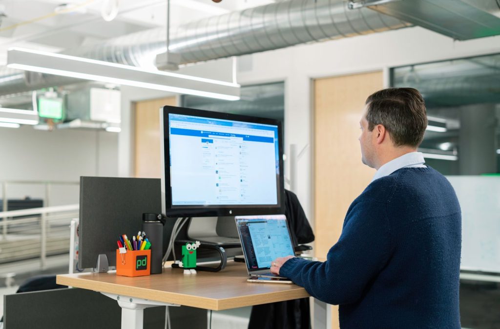 Homme travaillant debout devant un écran, à un bureau relevé dans un open space